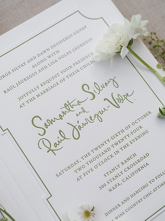 Wedding invitation with green wedding invitation calligraphy on deckle edge paper, framed by a white flower and greenery on linen fabric