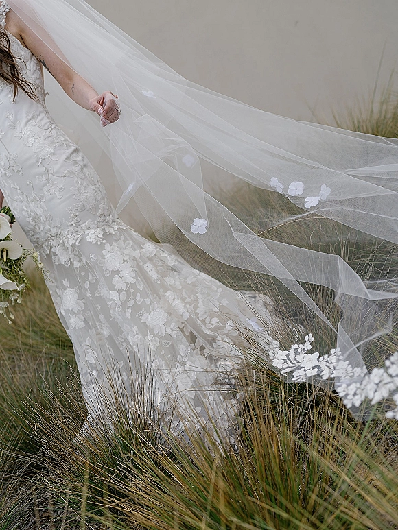 Bridal veil with cathedral length veil and 3D floral appliqué trailing behind bride holding calla lily bouquet by tall grass and neutral wall