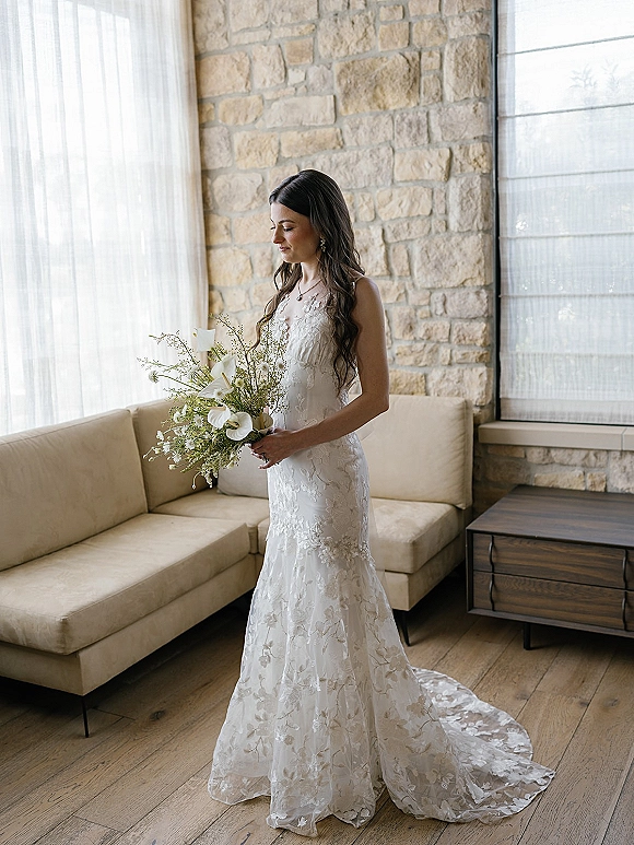 Bridal portrait of a bride holding bouquet of white calla lilies, looking down in a lace wedding dress by large windows and stone wall