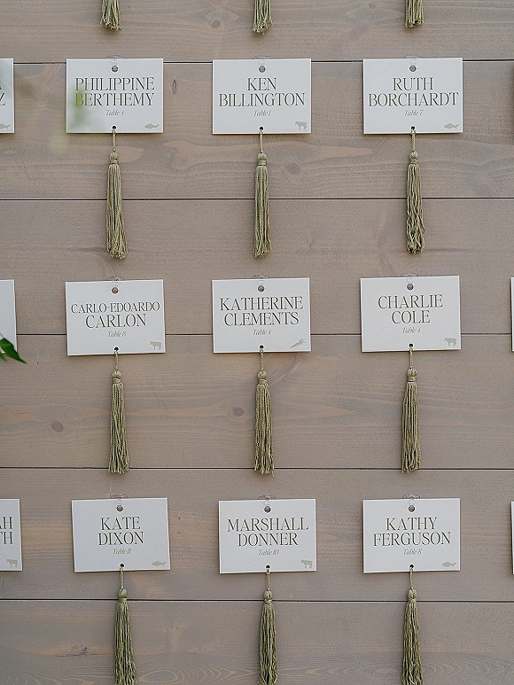 Seating chart display wedding seating chart with hanging escort cards, tassels, and clear clips arranged on a rustic wood panel backdrop
