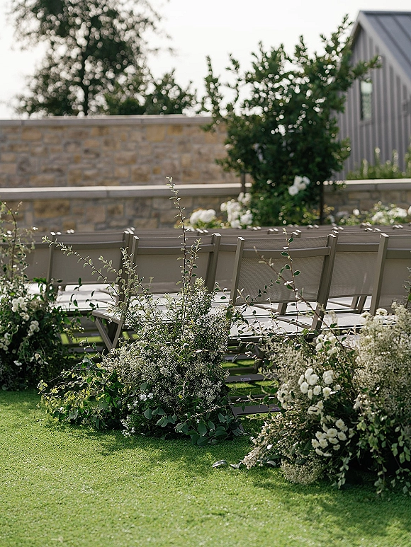 Ceremony seating with outdoor ceremony chairs lined on a lawn, aisle florals of white flowers and greenery near a stone wall backdrop