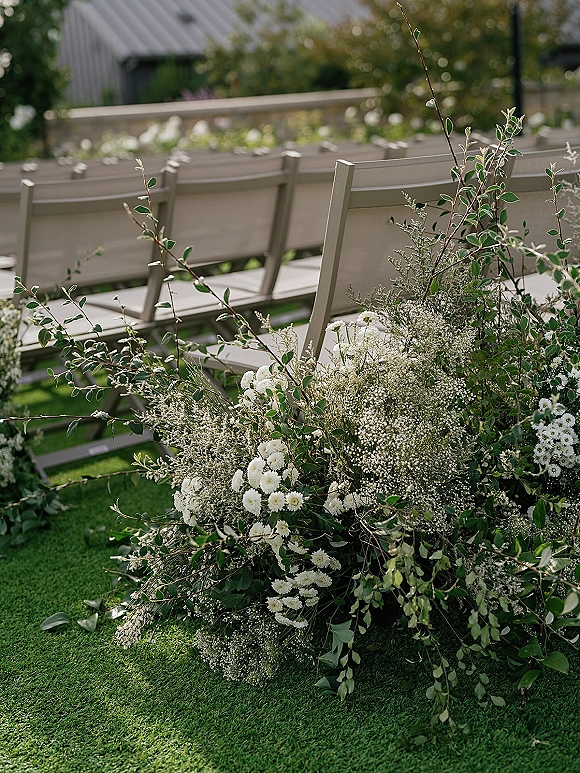 Ceremony aisle decor with outdoor ceremony aisle flowers, white blooms and baby's breath with greenery beside wooden chairs on a lawn