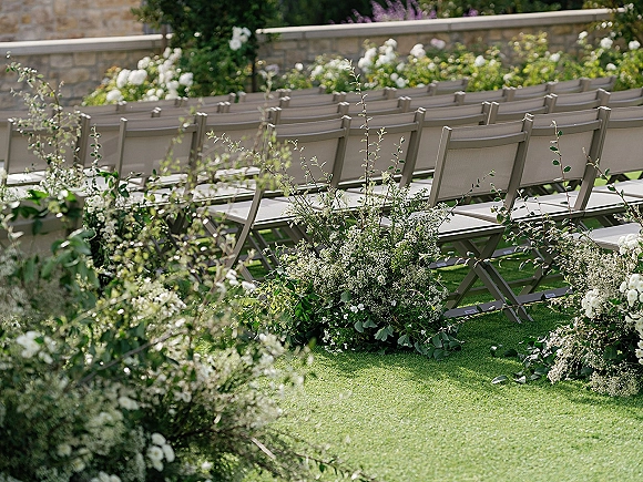 Ceremony seating with outdoor wedding ceremony chairs in neat rows, white florals and greenery lining the aisle on a grass lawn by a stone wall