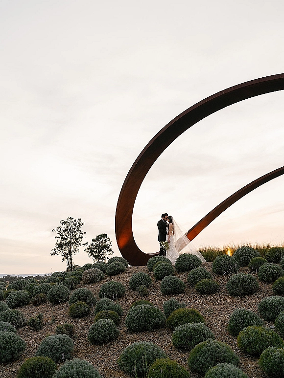 Wedding kiss portrait of bride and groom kiss beneath a sculptural metal arch, bride’s veil blowing as she holds a bouquet on a hillside