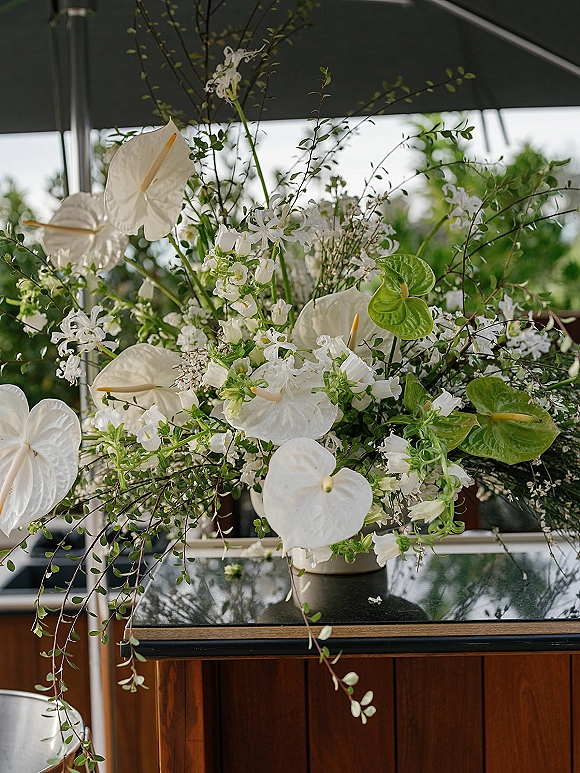 Wedding floral arrangement with white anthurium in a ceramic vase, accented by orchids and greenery on a table under an outdoor canopy