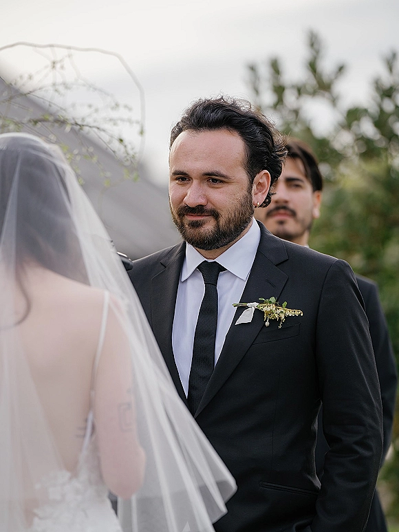 Ceremony moment as groom reacts at altar while bride approaches in veil and low-back dress, guests blurred amid outdoor greenery and sky