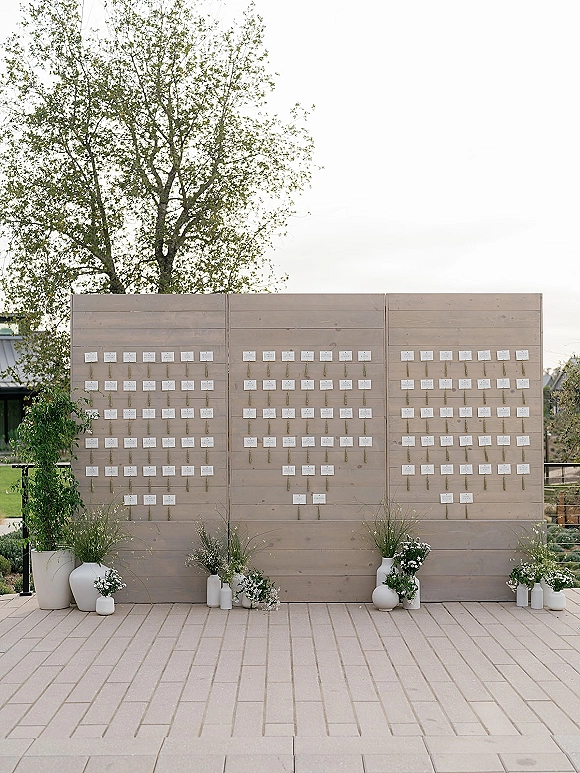 Wedding seating chart with escort card display on a wooden wall with brass hooks, flanked by white vases, greenery, and florals on an outdoor patio