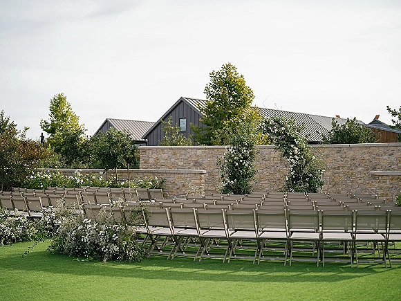 Outdoor ceremony setup with garden wedding ceremony chairs in a curved layout, facing a white flower and greenery arch on a lawn by a stone wall