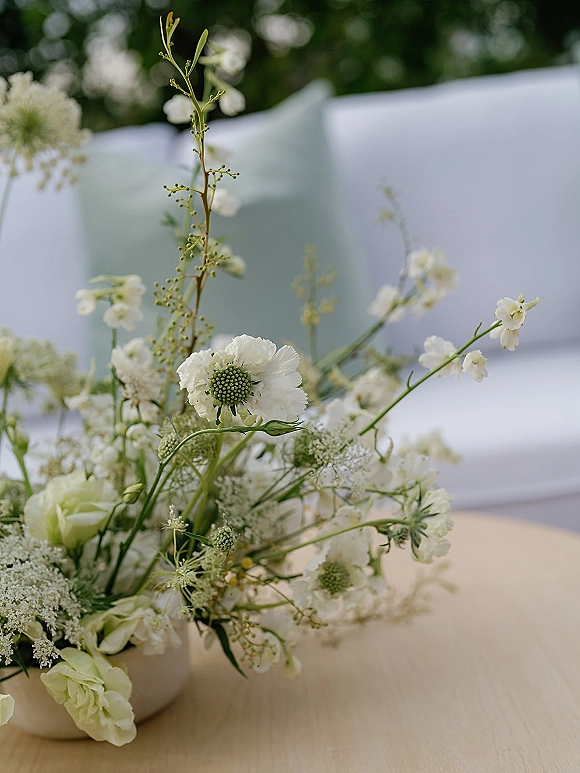 Wedding centerpiece with a white floral centerpiece in a ceramic vase on a wooden tabletop by a white couch and outdoor greenery