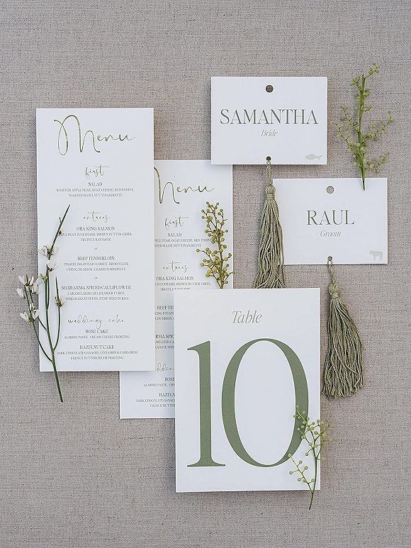 Wedding stationery flat lay with wedding menu card and place cards, tassel details and greenery sprigs on a linen fabric backdrop