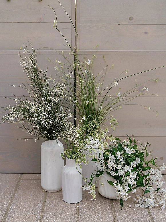 Wedding floral arrangement of white wedding florals in white ceramic vases with wildflowers and greenery against a wood plank wall