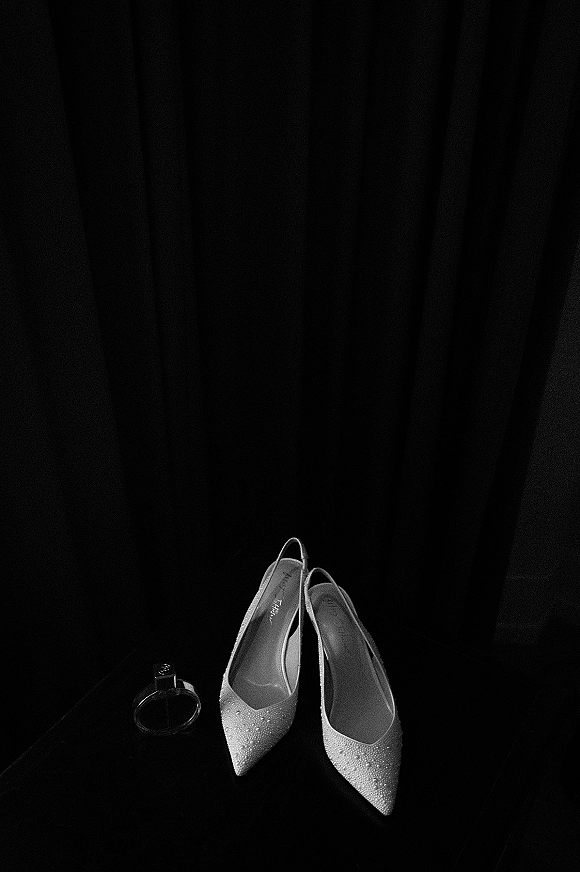Bridal shoes with rhinestone wedding heels set beside a perfume bottle on a dark surface against moody curtains backdrop