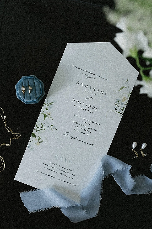 Wedding invitation suite with floral wedding invitation, RSVP card, velvet ring box and jewelry on a dark surface with blurred flowers