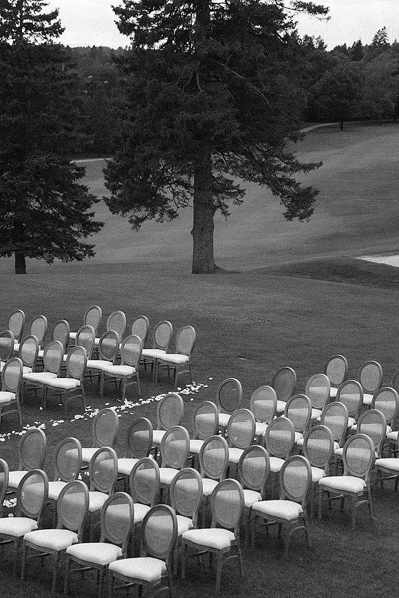 Outdoor ceremony setup with wedding ceremony chairs lined in rows and aisle petals scattered on grass, facing a tree-lined field backdrop