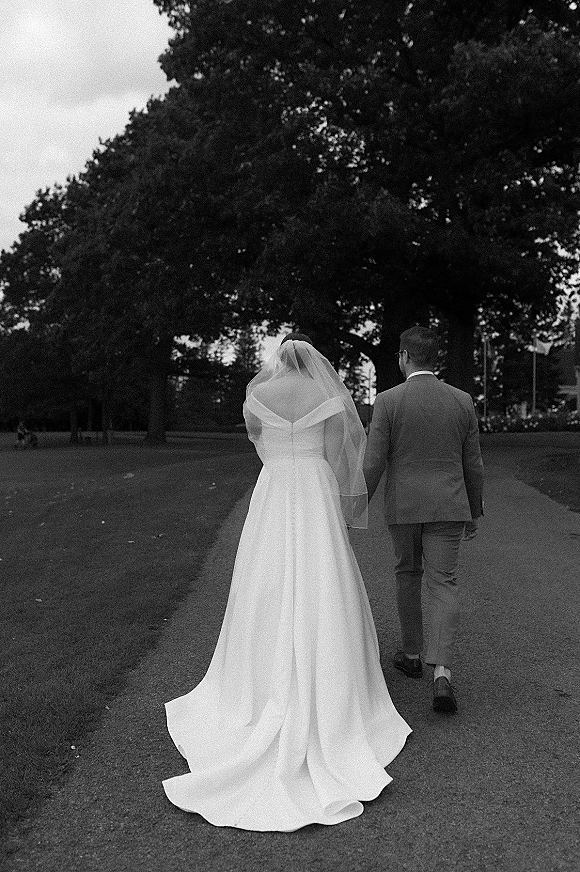 Couple portrait of bride and groom walking away hand in hand, her cathedral veil and dress train flowing down a tree-lined park path