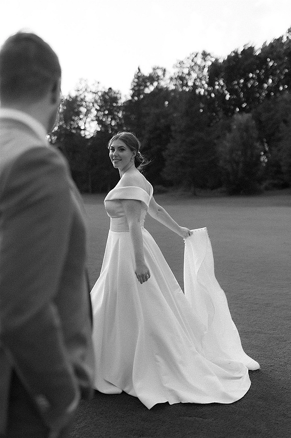 Bride portrait in an off the shoulder wedding dress with a long train and veil on a grassy lawn with trees and open sky