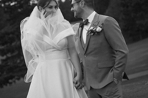 Couple portrait of bride and groom holding hands, her veil over face as they laugh on a lawn with trees behind them