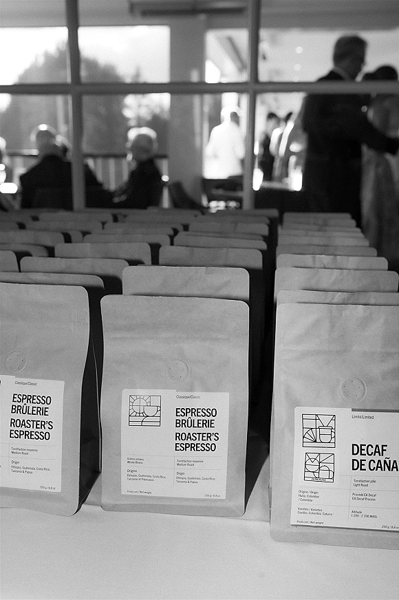Wedding favor table with coffee wedding favors in labeled bags on a tabletop, set by bright windows as guests mingle indoors