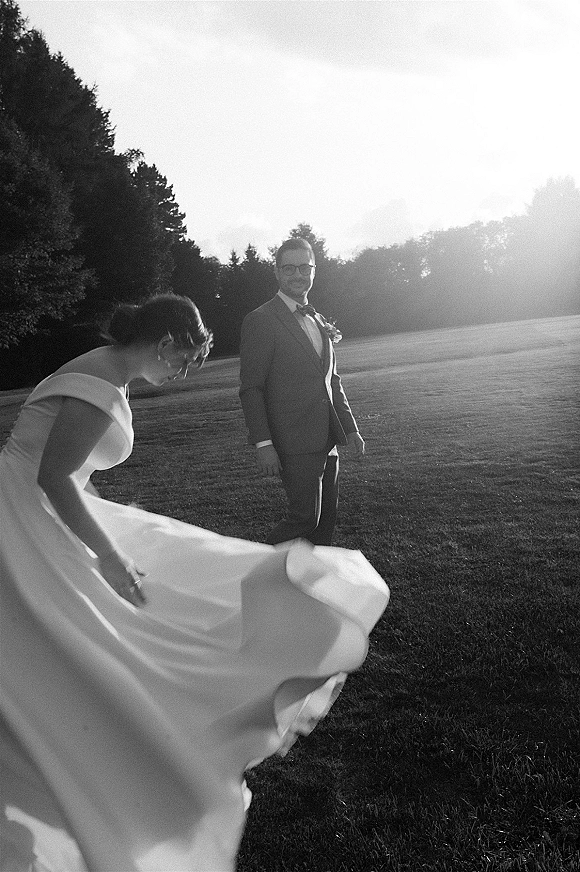 Couple portrait in a black and white wedding portrait, bride twirling her wedding dress beside groom in a bow tie on a sunlit lawn