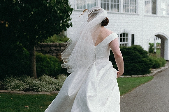 Bridal portrait of a bride walking away in a satin off-the-shoulder gown with chapel veil and low bun along a garden driveway