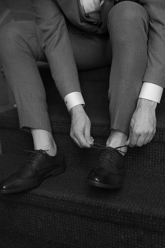 Groom getting ready, tying black dress shoes with suit pants and shirt cuffs on a carpeted floor beside a sofa indoors