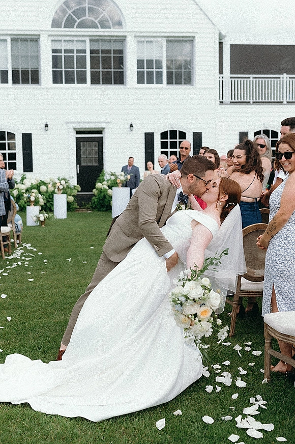 Wedding kiss as the groom dips the bride in her veil and off-the-shoulder dress, holding a bouquet, on a lawn by a white building