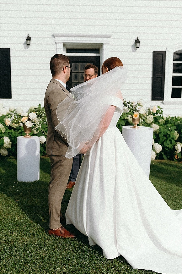 Wedding ceremony moment with bride and groom holding hands at the altar, veil trailing, beside white pedestal florals by a clapboard house