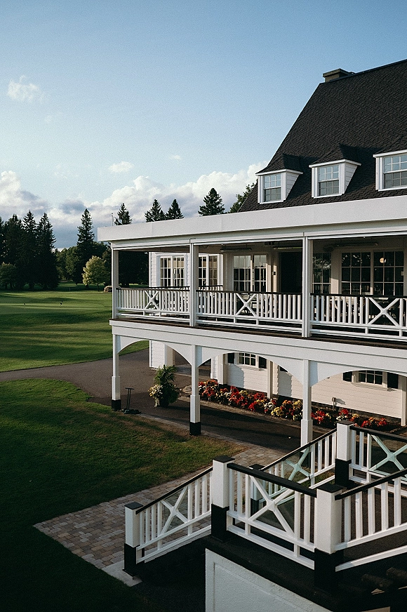 Wedding venue exterior with wraparound porch and white railings, dormer windows, and flower beds beside a golf course lawn and trees