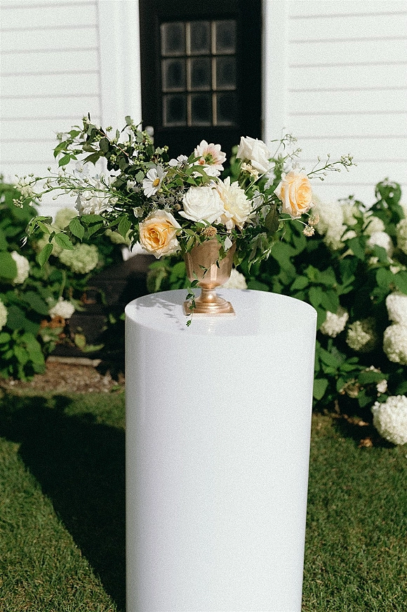 Wedding floral centerpiece in a gold compote centerpiece with garden roses, white blooms, and greenery on a white pedestal by hydrangeas