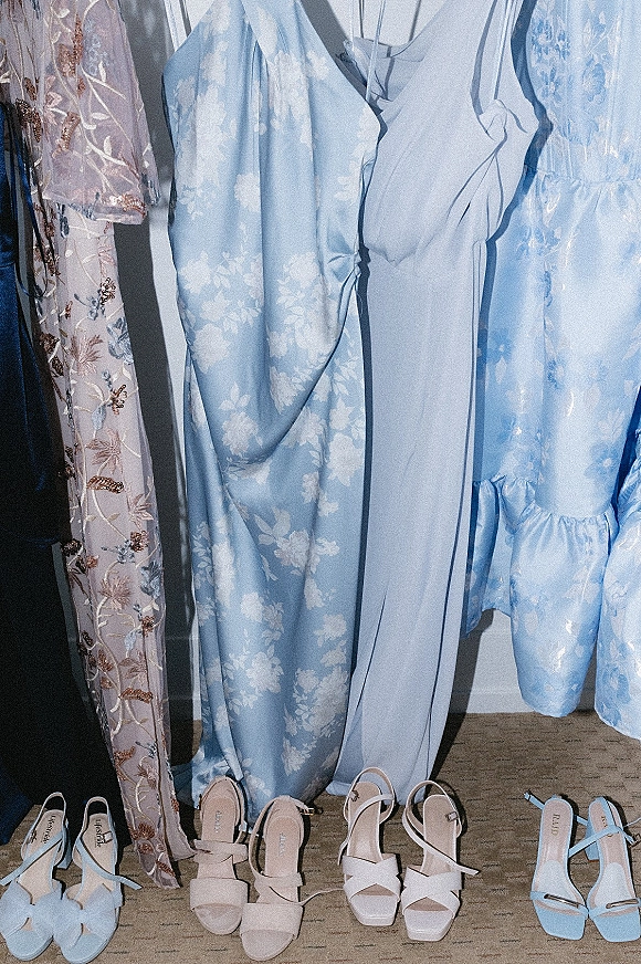 Bridesmaid dresses in pastel blue and floral prints hang in a closet, paired with strappy block-heel sandals on carpet floor