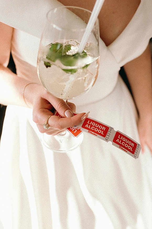 Wedding cocktail in a stemmed glass held by a bride in a white robe, engagement ring and manicure visible with drink tickets in soft light