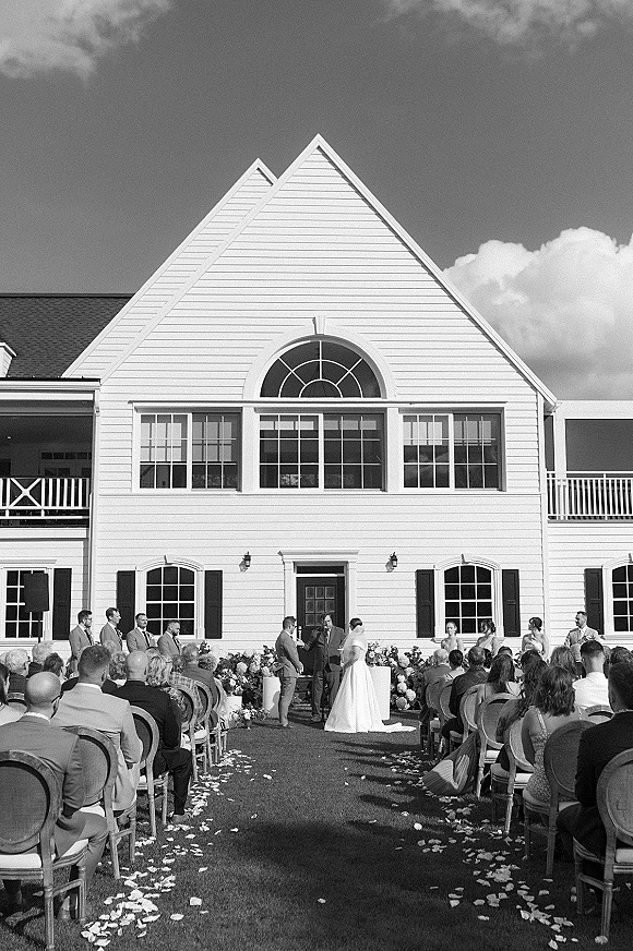 Outdoor wedding ceremony with ceremony aisle chairs strewn with rose petals, leading to a floral arch before a white house backdrop
