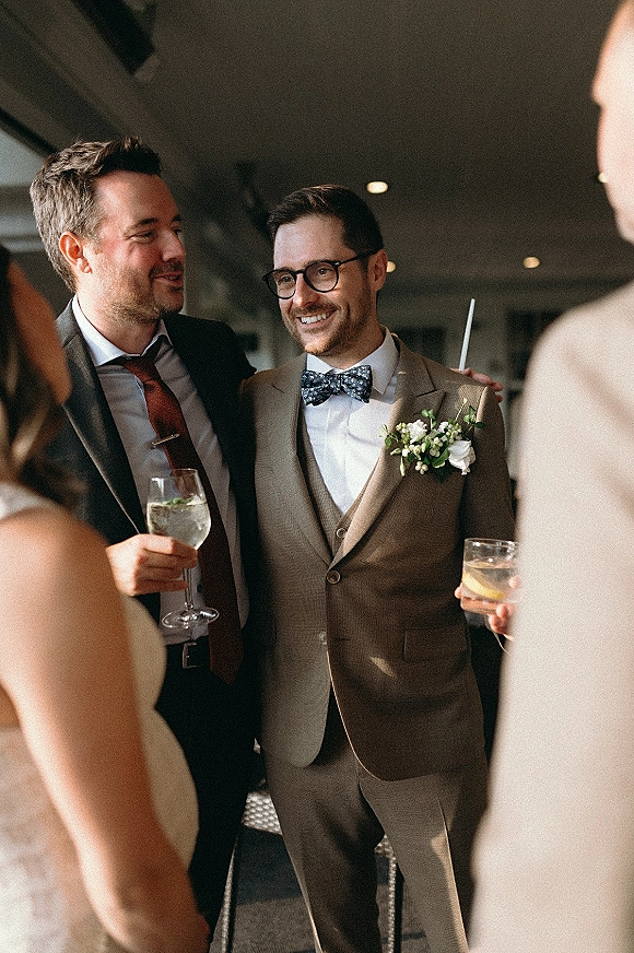 Groom with groomsmen in a groomsmen candid photo, laughing in tan suit with bow tie and boutonniere, holding drink glasses indoors under ceiling lights