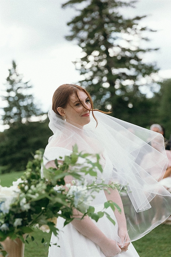 Bridal portrait of a bride in a bridal gown with a wedding veil blowing in the breeze, looking over her shoulder on a garden lawn