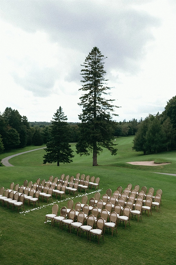 Outdoor ceremony setup with garden ceremony seating in a semicircle, cushioned chairs and white aisle petals on a golf course lawn under cloudy sky
