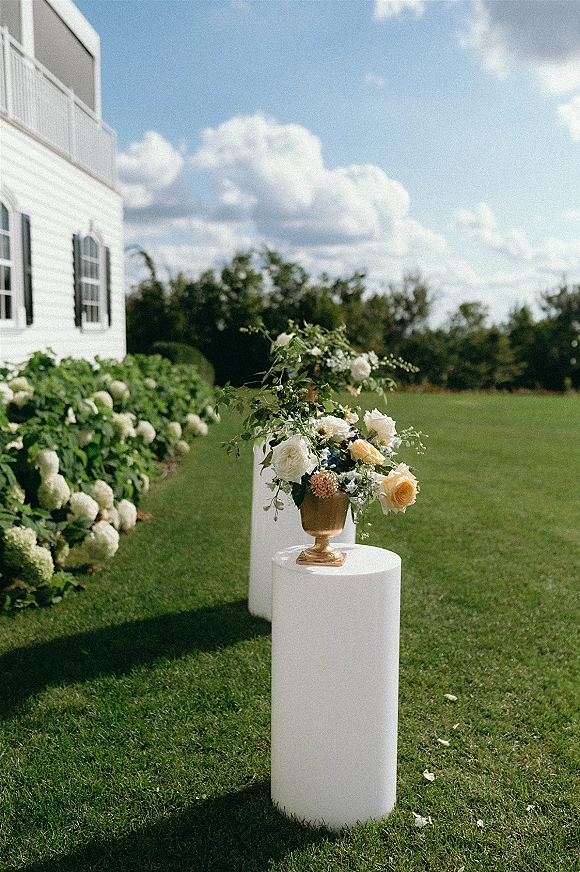 Ceremony floral arrangement in a gold urn on white pedestal plinths, with roses and greenery on a lawn beside a white house