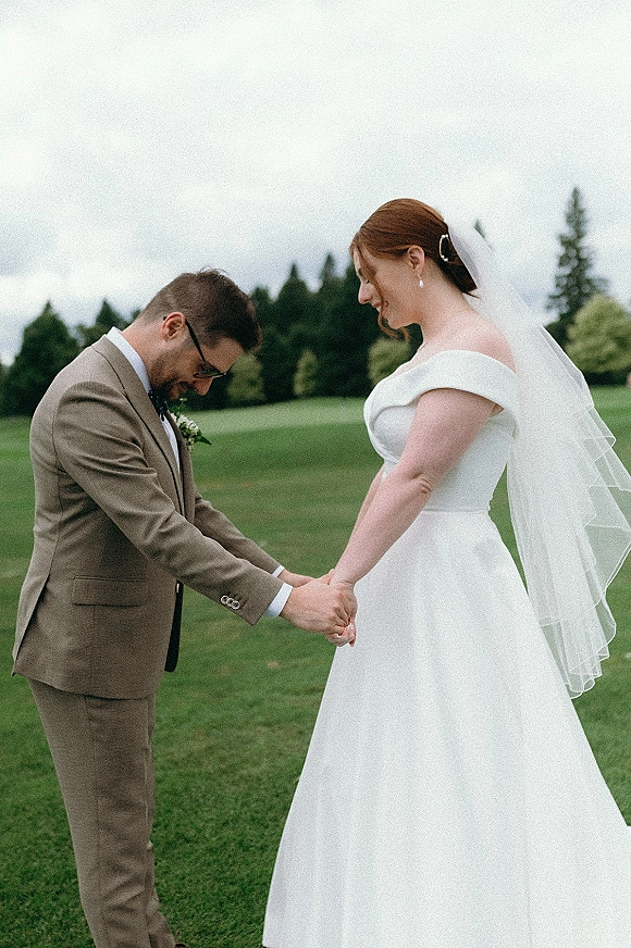 Wedding first look as bride in off-shoulder gown and veil holds hands with groom in glasses on a lawn beneath evergreen trees