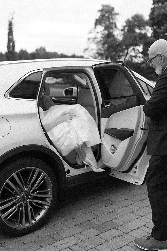 Bride arriving, stepping out of a car in high heels with veil and wedding dress as a suited man with eyeglasses assists on cobblestones under trees