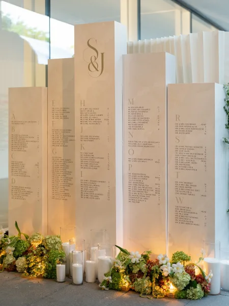 Wedding seating chart presented as a monogram seating chart against a glass wall with white drapery and a concrete floor.