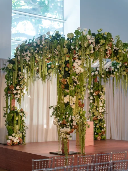Ceremony arch featuring hanging florals as indoor ceremony decor, framed by a large window with white curtains and a tree canopy beyond.