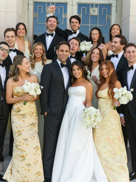 Wedding party portrait of newlyweds with their wedding party gathered on stone steps by a doorway against a stone facade, bouquets adding an accent.
