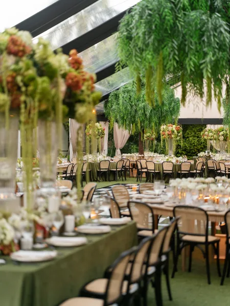 Reception decor with hanging greenery under a clear tent ceiling, long tables arranged among garden trees and manicured hedges.