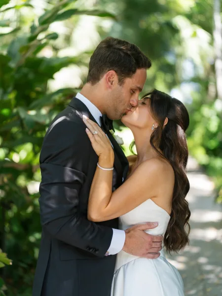 Wedding kiss portrait as the bride and groom kiss on a sunlit garden path, her diamond engagement ring catching light amid lush greenery.