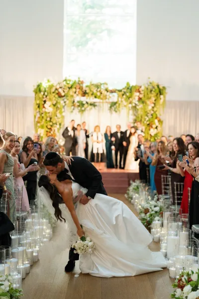 Wedding recessional kiss as a bride and groom dip, she clutching a white bridal bouquet, walking the ceremony aisle past cheering guests.