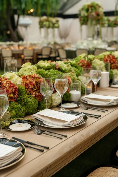 Reception tablescape for an outdoor reception table, hydrangeas lining a long setting beneath a tent canopy amid garden trees and chairs.