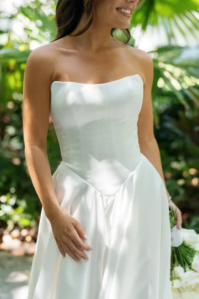 Wedding dress close-up of a strapless wedding dress as the bride holds a white bouquet amid palm-lined garden greenery.