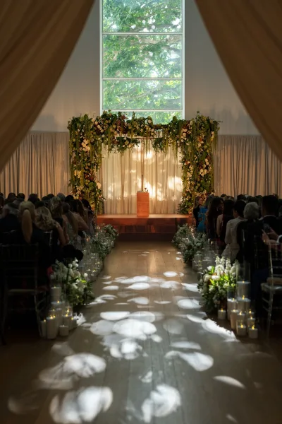 Ceremony setup showcasing indoor ceremony decor beneath a lush floral arch, set before a large window revealing a tree canopy.