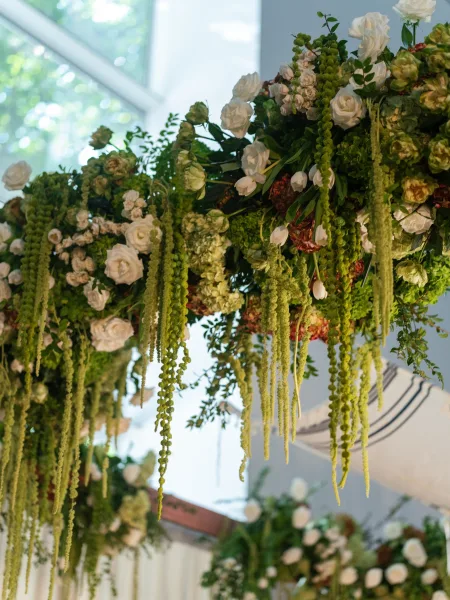 Ceremony arch flowers cascade as a hanging floral installation with green amaranthus under window light, indoors with garden trees beyond.