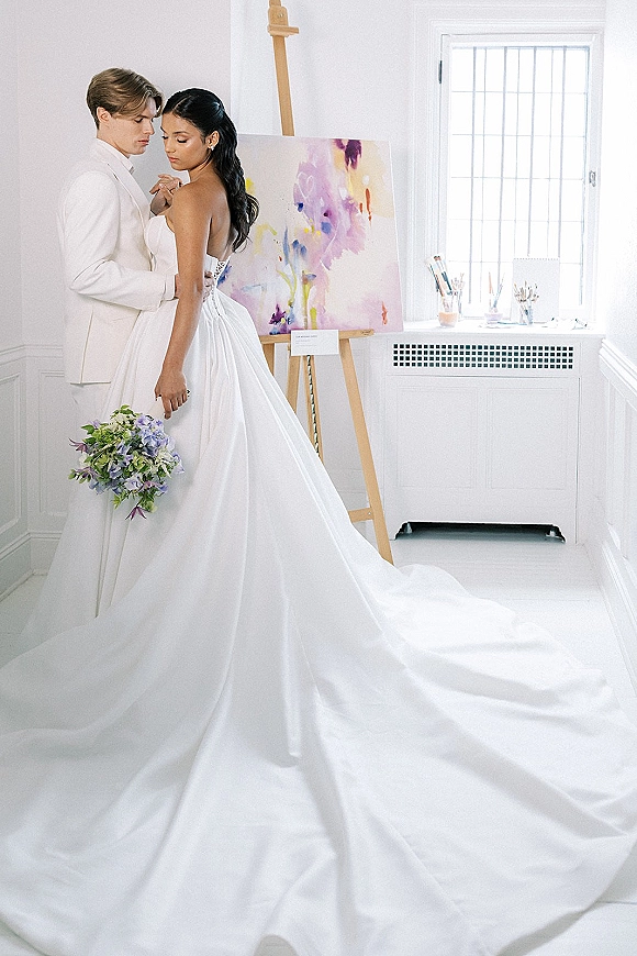 Couple portrait of bride and groom embrace, bride glancing over her shoulder in a strapless gown with long train by a window in a white room with an abstract painting on an easel