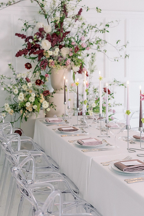 Reception tablescape wedding table setting with taper candles, blush napkins, glass chargers, coupe glasses, and florals against a white paneled wall